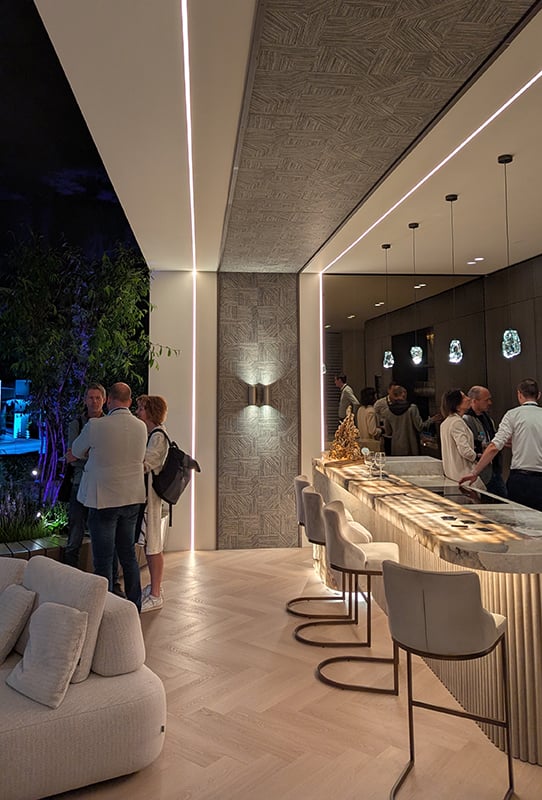 Detailfoto van de bar van Baas Architecten op The Art of Living beurs. Je kijkt op de kopse kant tegen de bar aan die een cremekleurig marmeren blad heeft met een soort golfplaat bekleding met krukken bekleed met lichte suede en donkerzwarte poten. Boven de bar hangen natuurstenen lampen met lichtdoorlatend kwartsiet. Ter hoogte van de bar een creme-kleurig wandpaneel met een brede bruine geometrische strip dat doorloopt in het plafond. Aan weerzijden van de strip een bijzondere COB-ledstrip.
