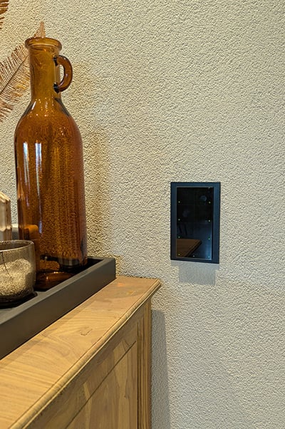 Een zwart custom-made wandpaneel van Grenton. Het verticale rechthoekige paneel bestaat uit 8 vlakken, opgedeeld in twee rijen van vier. In de buitenhoek van elk vierkant zie je een klein groen vierkant lichtje. Het paneel is bevestigd op een witte gespachtelde muur naast een eikenhouten dressoir. Op het dressoir staat een antraciet-grijze houten bak met daarin verschillende glazen vazen in bruin- en grijstinten.