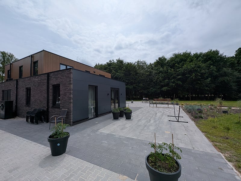 Moderne rechthoekige slimme villa in Drenthe. Begane grond bestaat uit rood-grijze bakstenen aan 1 zijde en donkergrijze platen aan de andere zijde met donkergrijs omlijste ramen en tuindeuren. 1e verdieping bestaat uit verticale roodkleurige houten planken. Deze verdieping is iets minder breed en lang. De tuin bestaat uit verschillende soorten en kleuren grijze tegels. Er staan grote donkergrijze potten met planten her en der, evenals grijze tuinstoelen en een houten bank. Verder een badmintonnet. Om de tuin gras en bomen.
