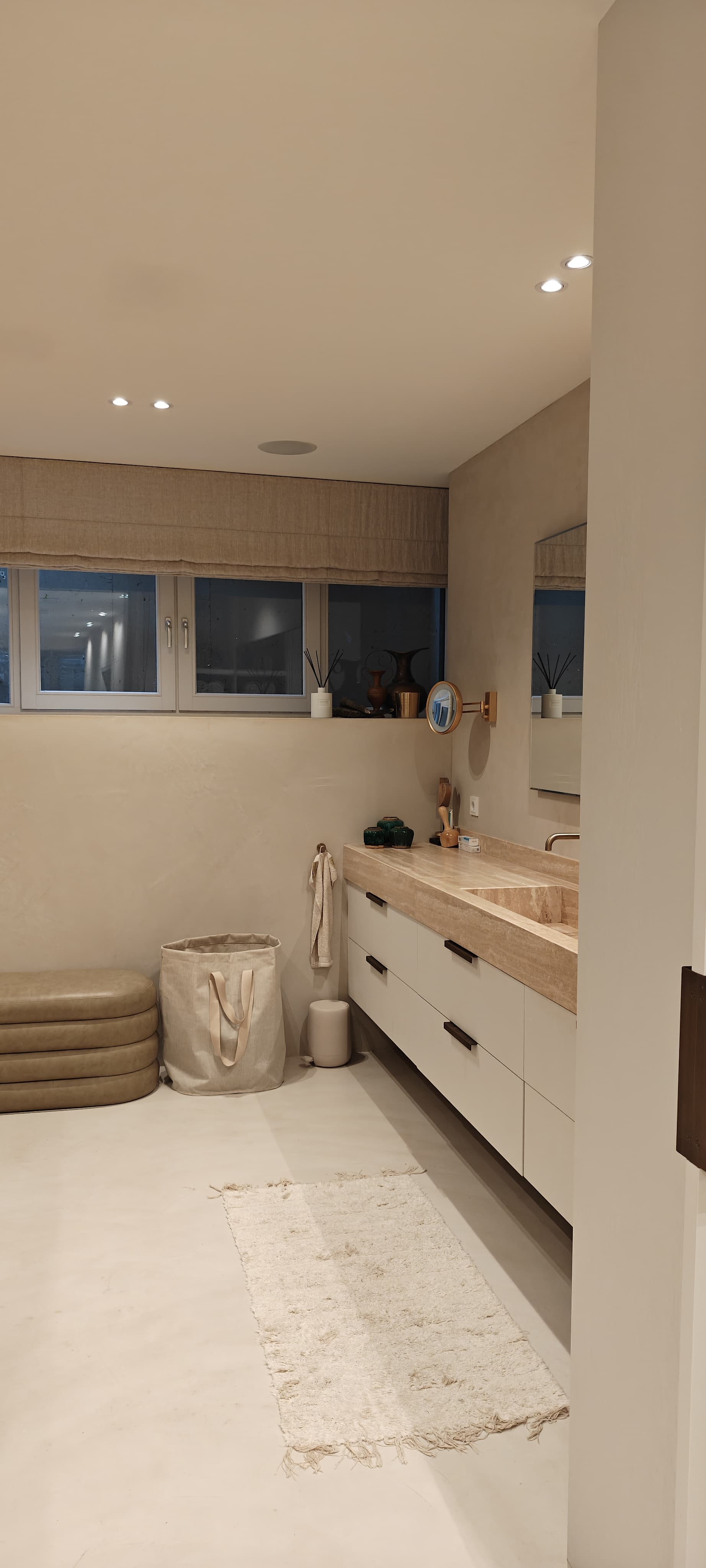 Badkamer in souterrain van Amsterdams pijpenla appartement. Ramen zitten hoog en komen zichtbaar niet boven straat uit. Badkamer is in beige tinten geschilderd met elektrisch beige vouwgordijn. Rechts zie je een beige marmeren badkamerblad met een spiegel erboven en een cremekleurige ladenkast eronder.