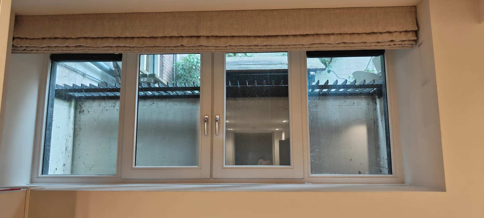 Detailfoto raampartij in slaapkamer souterrain Amsterdams pijpenla appartement. Raam bestaat uit vier delen en grijze sponningen. Middelste twee ramen kunnen open. Voor het raam zit een beige elekrisch schuifgordijn. Muren en kasten zijn licht beige. In het plafond zitten links voor de kastenwand spotjes.