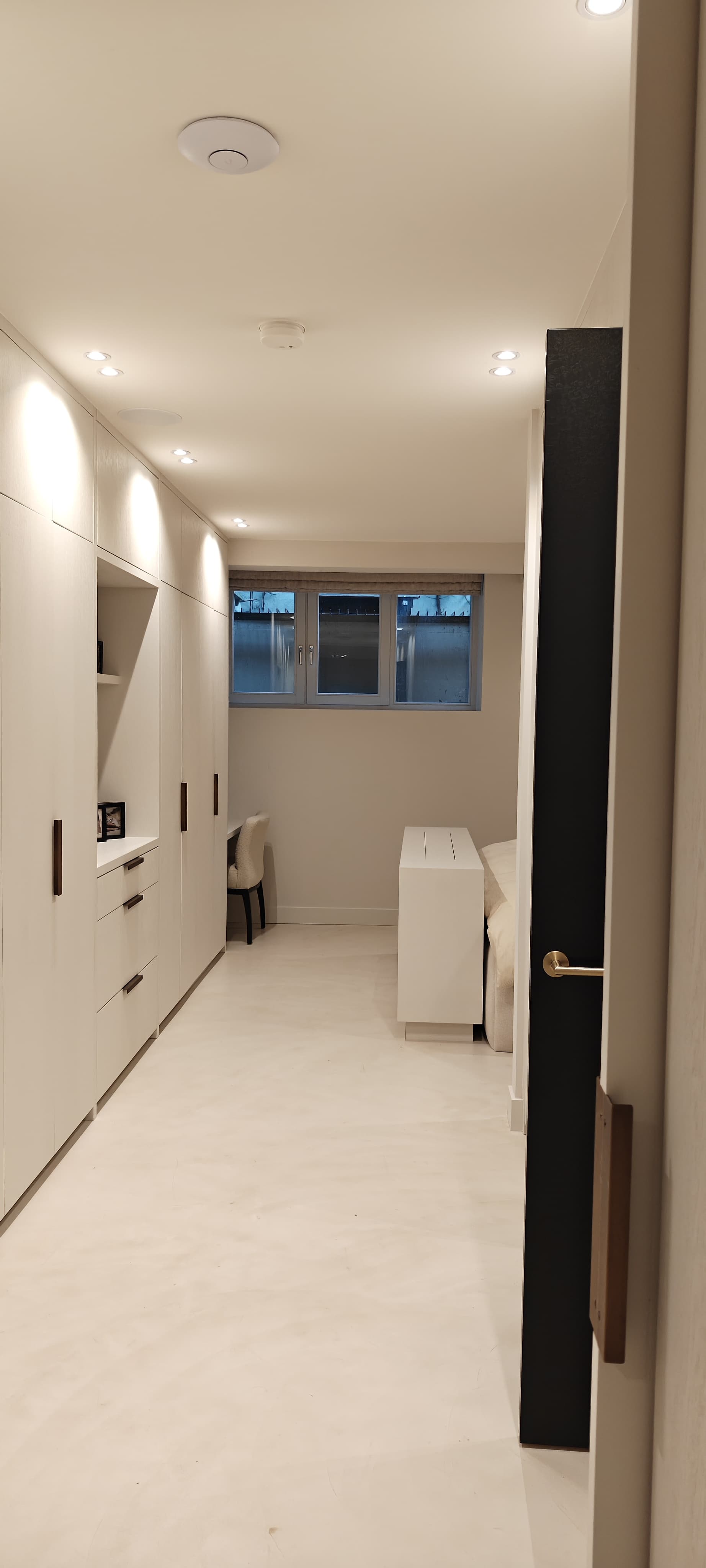 Slaapkamer Amsterdamse pijpenla, souterrain te zien aan het raam dat uitkomt onder de straat. Kamer is gebroken wit geschilderd. Evenals de plafondhoge kastenwand links. Rechts zie je nog net het uiteinde van een bed met aan het voeteneinde een hocker waarin een tv zit verwerkt die omhoog kan komen. In het plafond meerdere spotjes. In de linkerhoek naast de kastenwand zie je nog net een klein bureautje met beige stoel ervoor.