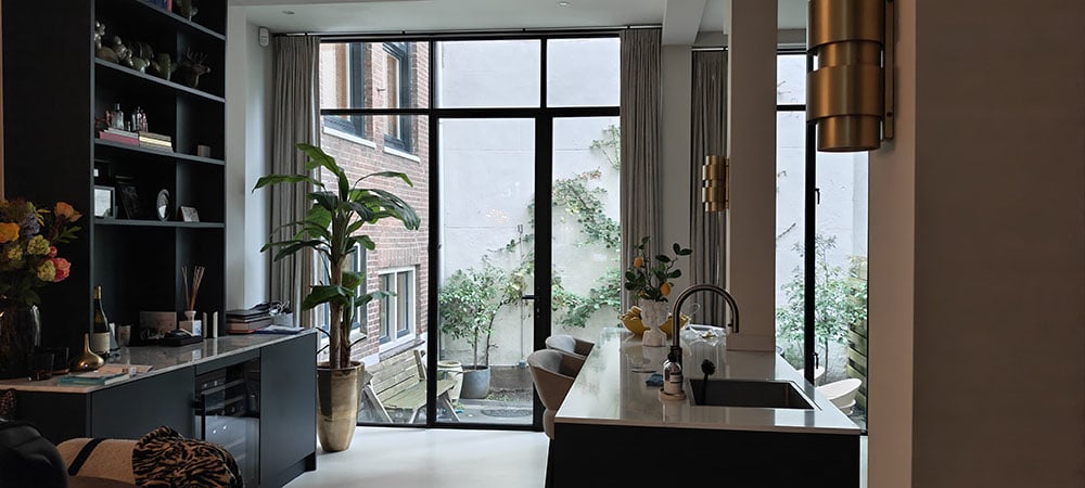 Moderne zwarte keuken in amsterdamse pijpenla. Kasten matzwart. Links een aanrecht met beige marmer en een wijnkoelkast. Plafondhoge ramen met uitkijk op kleine binnentuin. In linkerhoek een grote groene kamerplant. Boven het aanrecht een open zwarte kast met planken met daarop snuisterijen. Rechts een keukeneiland met beige aanrechtblad met zwarte vierkante gootsteen met koperen kraan. Aan de linkerkant van het keukeneiland twee beige stoelen. Rechts zie je een beige pilaar met koperen wandlamp. Op de achtergrond plafondhoge ramen met glazen deur naar binnentuintje.