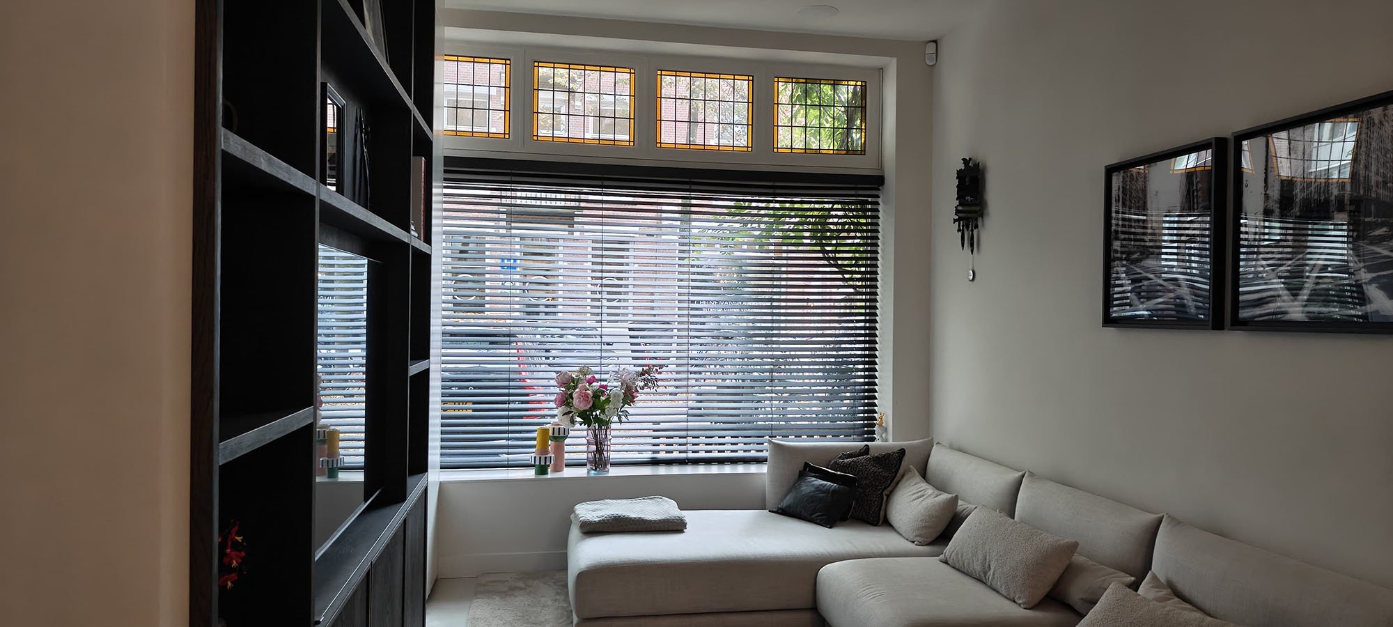 Woonkamer Amsterdams pijpenla appartement. Je kijkt uit op de straat. Met 4 glas-in-lood raampjes aan bovenzijde voorraam. Zwarte luxaflex. Links zie je nog net een plafondhoge open zwarte tv kast. Rechts tegen de beige muur een grote licht grijs-beige hoekbank. In het raamkozijn een vaas met bloemen en twee kleurige kandelaars met kaarsen.