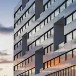 Gevel van University Hospital in Geneve. Zon gaat onder en de oranje gloed weerkaatst in de ramen van het gebouw. Het gebouw bestaat uit 9 verdiepingen, met grijze panelen afgewisseld met ramen met zwarte sponningen. Deel van het EU project INBLANC duurzaam wonen en werken waaraan Comfortica deelneemt.