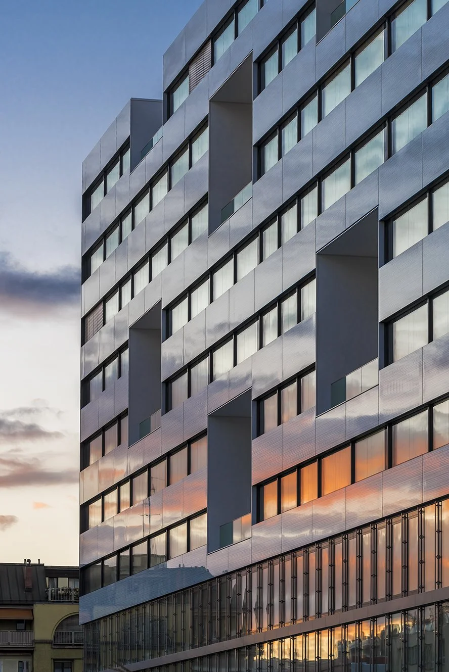 Gevel van University Hospital in Geneve. Zon gaat onder en de oranje gloed weerkaatst in de ramen van het gebouw. Het gebouw bestaat uit 9 verdiepingen, met grijze panelen afgewisseld met ramen met zwarte sponningen. Deel van het EU project INBLANC duurzaam wonen en werken waaraan Comfortica deelneemt.