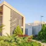 Zandkleurig publiek gebouw omringd door groen in de stad Thessaloniki in Griekenland. De gevel bestaat uit lichte houtkleurige horizontale planken. Op de achtergrond lichtgrijze gebouwen met lichtgrijze planken. Deel van het EU project INBLANC duurzaam wonen en werken waaraan Comfortica deelneemt.