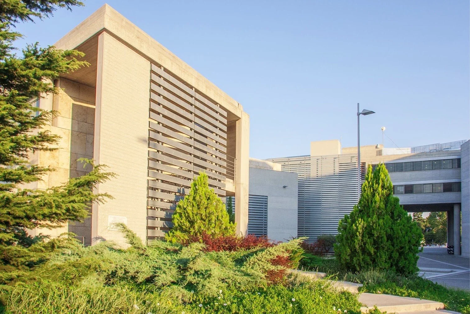 Zandkleurig publiek gebouw omringd door groen in de stad Thessaloniki in Griekenland. De gevel bestaat uit lichte houtkleurige horizontale planken. Op de achtergrond lichtgrijze gebouwen met lichtgrijze planken. Deel van het EU project INBLANC duurzaam wonen en werken waaraan Comfortica deelneemt.