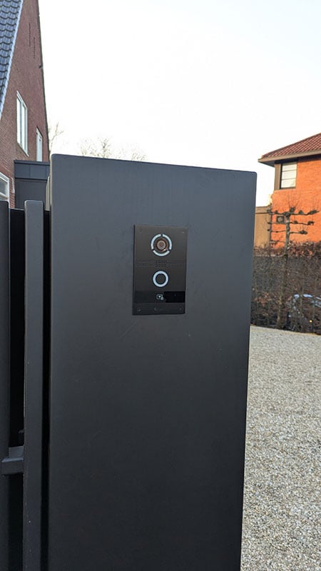 Zwart dicht hekwerk met daarop een zwart intercom-paneel met camera en bel. Je ziet rechts een grote oprit met grind. Daarnaast een haag en de hoek van een rood bakstenen huis met laag schuin dak met rode dakpannen. Links zie je nog net een puntdak van een bruin-rood stenen huis met zwarte dakpannen.
