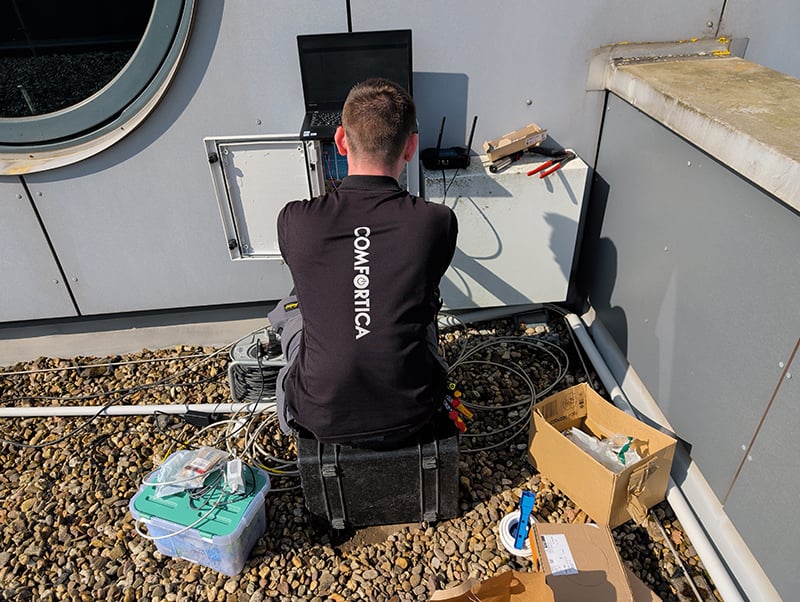 Monteur met kort donkerbruin haar en zwart t-shirt met Comfortica logo zit op het platte dak van 3L Architekten in Duitsland. Je ziet hem op zijn rug terwijl hij op een zwarte werkkoffer zit en bezig is met elektriciteitskasten. Om hem heen zie je dozen en bedrading liggen en een laptop.
