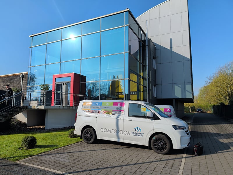Twee witte bedrijfsbusjes met de logo's van Comfortica en Slimmer Wonen voor het pand van 3L Architekten. Voorste gebouw is van glas met vierkante sponningen. Rode rand om dubbele voordeur. Er schuinachter tegen staat een iets hoger grijs gebouw met matgrijze rechtopstaande panels met afgerond dak.