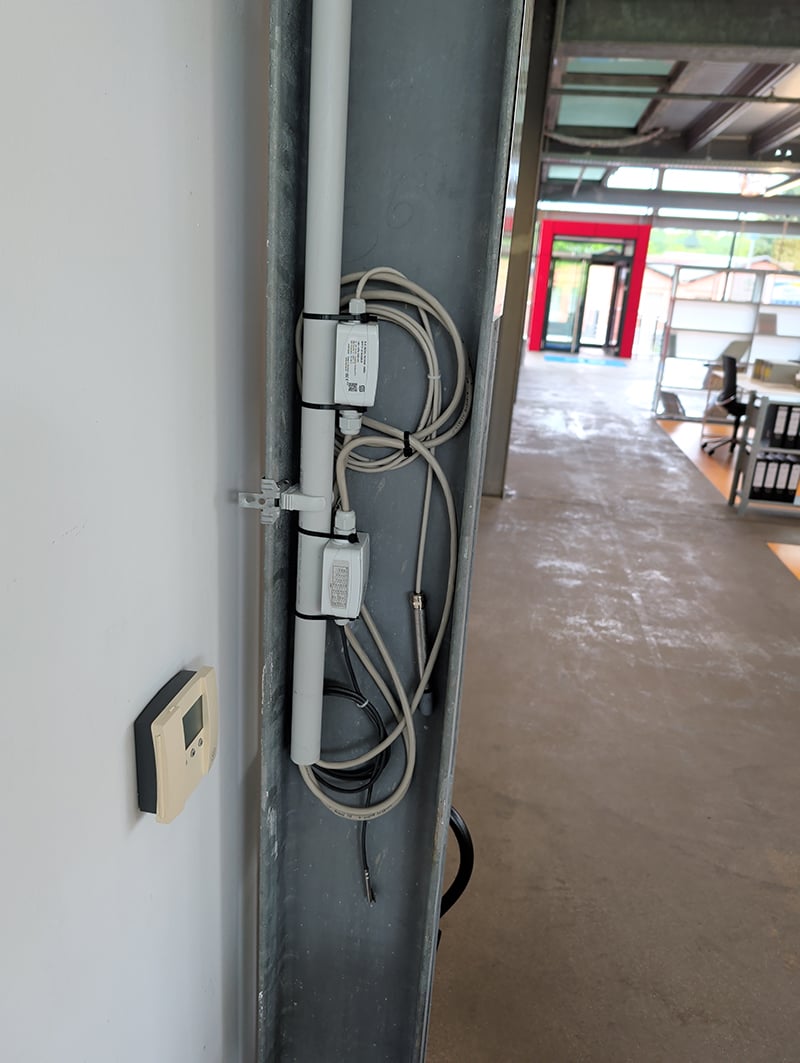Sensoren voor duurzaamheidsmetingen bij 3L Architekten. Je ziet een stalen frame tegen een grijze muur. Tegen de linker binnenhoek zit een grijze buis waaruit grijze en zwarte elektriciteitsdraden komen die geleid worden door grijze doosjes die tegen de grijze buis zitten. Rechts kun je het kantoor in kijken. Je ziet een betonnen vloer en op de achtergrond de glazen voorpui met dubbele voordeur.