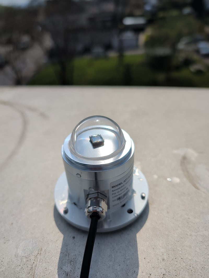 Sensor op het dak van 3L Architekten die onder andere wind meet. Je ziet een aluminium rond statief die met bouten aan de betonnen dakrand is vast gezet. Op het statiefje zit een vierkant zwart sensortje met een doorzichtig koepeltje eroverheen.