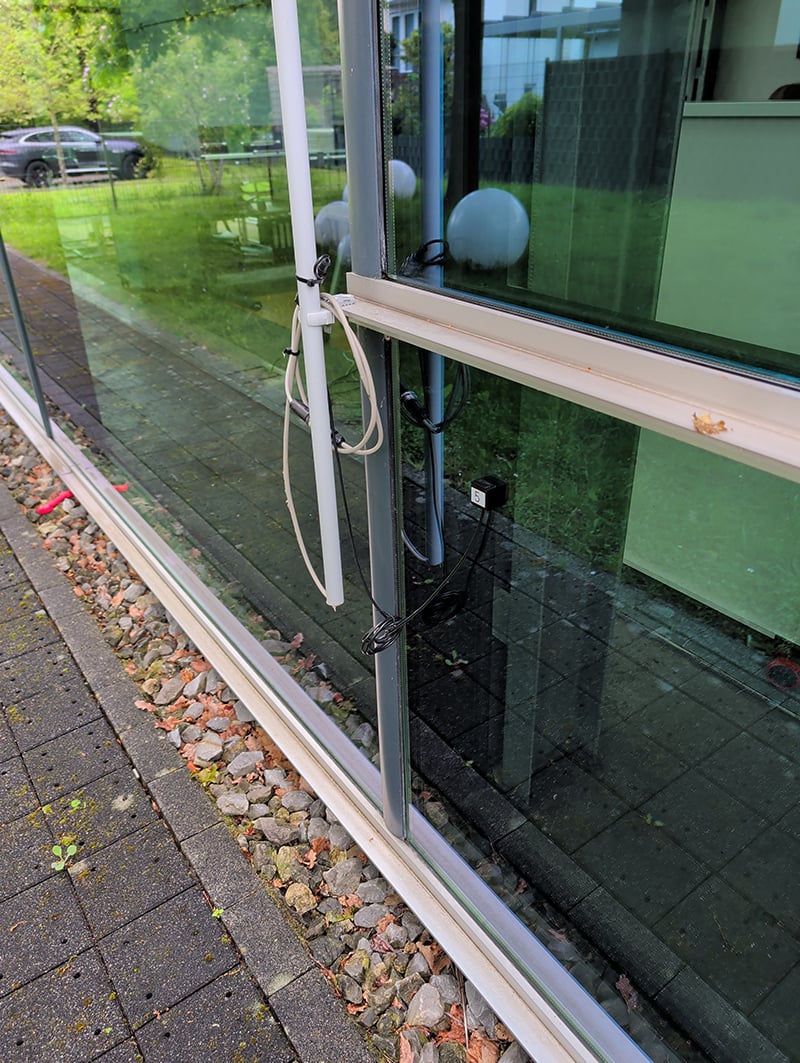 Buitenzijde glazen pui bedrijfswand waar langs een grijze buis loopt met bedrading en sensor die op het glas is bevestigd. In de glazen pui zie je het groene gazon en bomen weerspiegeld.