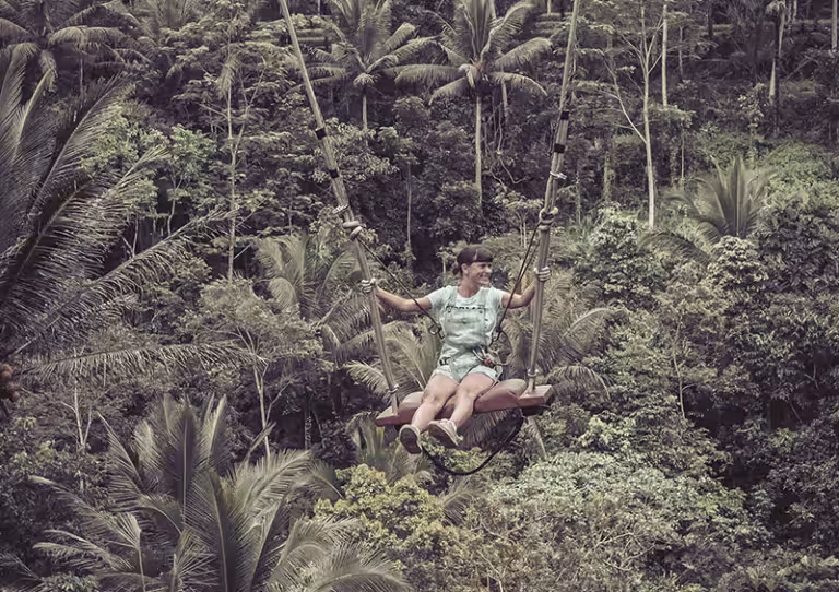 Op de achtergrond een enorme jungle. Op de voorgrond een jonge vrouw op een hele lange schommel die schijnbaar boven de jungle schommelt. Ze heeft een afgeknipte lichtblauwe spijkertuinbroek aan, een wit t-shirt, blote voeten en donker haar in twee knotjes aan de zijkant van haar hoofd. Ze kijkt heel blij naar rechts. Ze houdt met haar beide handen de schommeltouwen stevig vast.